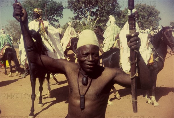 Celebrating the end of Ramadan. A barechested Nigerian man poses for the camera, waving two sticks in the air at a celebration to mark the end of Ramadan. Zaria, Nigeria, 1958. Zaria, Kaduna, Nigeria, Western Africa, Africa.