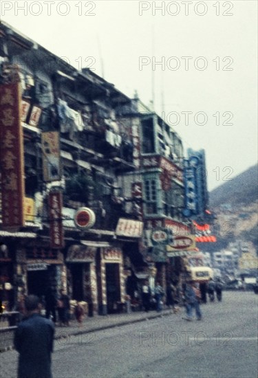 Street scene in Kowloon. Shopfronts on a busy city street are littered with signs written in Chinese script, some illuminated by neon light. Kowloon, People's Republic of China, March 1960. Kowloon, Hong Kong, China, People's Republic of, Eastern Asia, Asia.