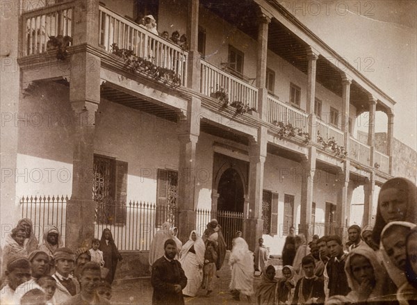 Russi's house' in Safi. Crowds of people stare in wonderment at the camera beside a large colonial-style building identified as 'Russi's House'. The building has two storeys, a long wooden balcony running across the first floor and shuttered windows. Safi, Morroco, 1898. Safi, Safi, Morocco, Northern Africa, Africa.