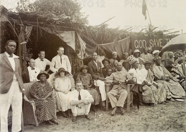 Empire Day celebrations. Informal group portrait of Africans and Europeans at Empire Day celebrations at Badragry. The group includes European employees of Miller Brothers and the African Oil Nuts Company and an African priest, probably an Anglican. A partially obscured banner reads 'God Save the King'. Nigeria, May 1924. Nigeria, Western Africa, Africa.