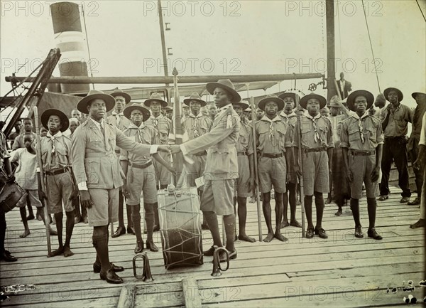 Boy scouts. Change of watch or duty between two Nigeria Boy Scout leaders onboard a steamer. The two Scout masters shake hands across a large kettle drum that stands between them, watched by an audience of at least 20 African Boy Scouts who are all standing to attention. Nigeria, 1928. Nigeria, Western Africa, Africa.