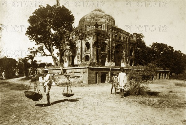 Ruined mosque, Serampore. A man carries baskets on a pole over his shoulder at the site of a ruined mosque. The stonework of the mosque is crumbling, the walls overgrown with vegetation. Serampore, India, circa 1890. Serampore, West Bengal, India, Southern Asia, Asia.