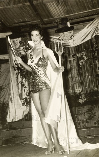 Carnival Queen beauty pageant. A beauty pageant contestant, Wendy Lawrence, poses on stage holding a trident in one hand and a wand in the other. She is dressed in a sequinned leotard and a long cape, and wears a sash advertising 'Carr's Biscuits'. Grenada, 2 March 1957. Grenada, Caribbean, North America .