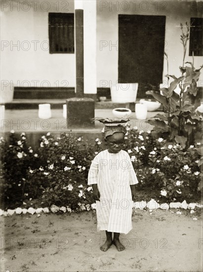 Young boy, Nigeria. A young African boy, identified as 'Bobby', is pictured in the garden of a bungalow wearing a simple striped robe as he balances a plate on his head. Nigeria, circa 1925. Nigeria, Western Africa, Africa.