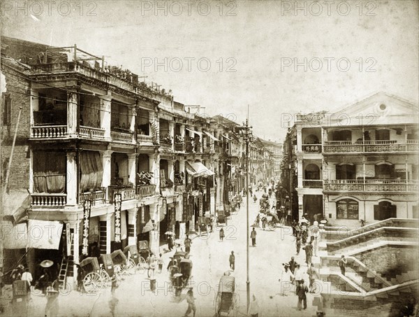 Street scene in Hong Kong. Pedestrians and rickshaws travel along a busy street in Hong Kong. Colonial-style houses with balconies line both sides of the street. Hong Kong, (People's Republic of China), circa 1895. Hong Kong, Hong Kong, China, People's Republic of, Eastern Asia, Asia.