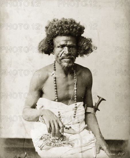 Portrait of an ageing man. The man is shown seated, with a cloth wrapped around his chest and lower body. A dagger is held in a holster attached to his left elbow. In his right hand he holds what appears to be a charm. He has a long string of beads, possibly seeds, around his neck. North Africa, circa 1885., Northern Africa, Africa.