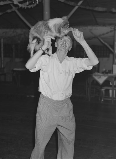 Tom dances with a dog. A man identified as 'Tom', lifts a worried-looking collie dog onto his shoulder at a Christmas party being held at the Forest Inn bar. Possibly Nairobi, Kenya, 28 December 1952. Kenya, Eastern Africa, Africa.