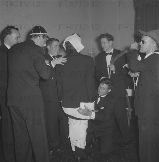 Rugby team antics. Members of a rugby team dressed in tuxedos fool around at the Mogambo Club. Two of the group attempt to 'mummify' their friend by wrapping hm in toilet paper. Possibly Nairobi, Kenya, 21 September 1957. Kenya, Eastern Africa, Africa.