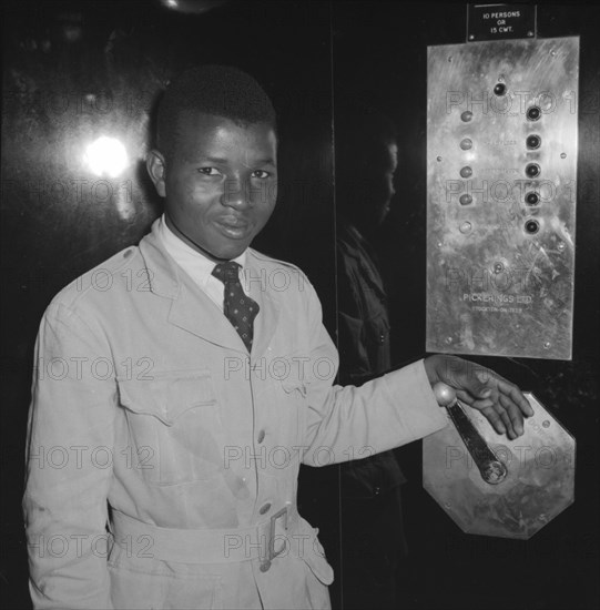 Congress House lift attendant. A young African lift attendant smiles for the camera, his hand resting on a manual handle inside an elevator at Congress House. Nairobi, Kenya, 11 October 1957. Kenya, Eastern Africa, Africa.