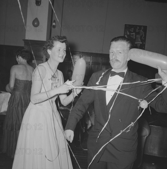 Single balloon salute. A middle-aged man at a Northern Counties dance, salutes the camera with a balloon as his partner looks on, amused. Kenya, 2 November 1957. Kenya, Eastern Africa, Africa.