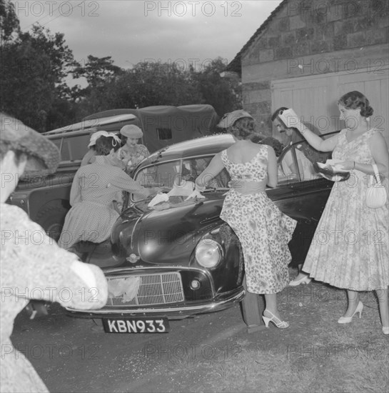 Wedding send off. Female wedding guests surround a car, excitedly throwing confetti over the newlywed Coltart couple as they climb into the back seat. Kenya, 9 November 1957. Kenya, Eastern Africa, Africa.