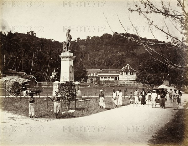 Statue of Sir Henry Ward. Statue of Sir Henry Ward (1797-1860), Governor of Ceylon between 1855 and 1860, in front of the Dalada Maligawa or Temple of the Buddha's Tooth. Several people walk along the road beside the statue, some shaded by umbrellas. Kandy, Ceylon (Sri Lanka), circa 1875. Kandy, Central (Sri Lanka), Sri Lanka, Southern Asia, Asia.