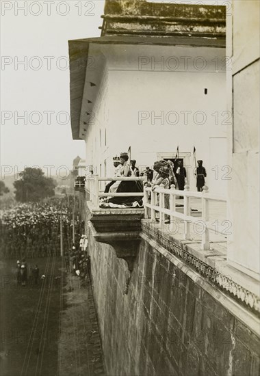 The day after the Coronation Durbar. King George V (r.1910-36) and Queen Mary appear before crowds from a throned balcony at the Delhi Fort. Pictured the day after the Coronation Durbar, they are dressed in their coronation robes and crowns, and are accompanied by ten Indian pages. Delhi, India, 13 December 1911. Delhi, Delhi, India, Southern Asia, Asia.