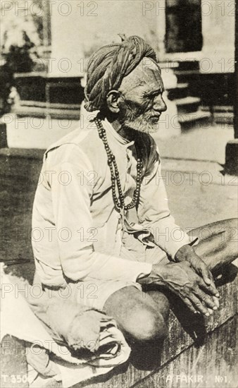 An elderly fakir. Portrait of an elderly fakir sitting cross-legged on a wall. Dressed in a short kaftan that exposes his knees, he wears a turban and a string of beads around his neck. India, circa 1910. India, Southern Asia, Asia.