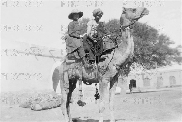 Miss Arthur rides a camel. A European lady identified as Miss Arthur sits on a camel behind a 'shikari' (professional hunter), ready to embark on a hunting expedition to the countryside. Probably Sind, India (Sindh, Pakistan), December 1911., Sindh, Pakistan, Southern Asia, Asia.