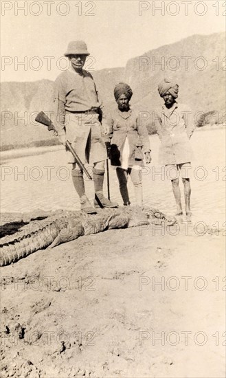 Shot by the Acting Governor of Bombay. Sir Henry Staveley Lawrence, Acting Governor of Bombay, poses rifle in hand as he stands on the carcass of a crocodile he has shot. He is pictured on a riverbank, accompanied by two Indian 'shikaris' (professional hunters). Deoli, Bombay Presidency (Maharashtra), India, circa 1926. Deoli, Maharashtra, India, Southern Asia, Asia.