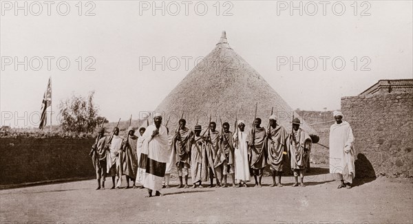 Armed guard at the Sudan-Ethiopia border. A number of armed Ethiopian men stand in front of a round, thatched hut at Gallabat, guarding the border crossing between Sudan and Ethiopia. A union jack flag is raised on a flagpole to the left of the photograph. Gallabat, Sudan, circa 1910. Gallabat, Al Qadarif, Sudan, Eastern Africa, Africa.