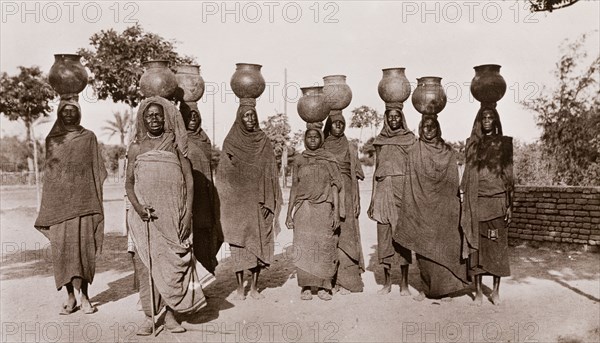 Sudanese women carrying water. A group of Sudanese women, dressed in long robes and headscarves, walk along a road carrying water in ceramic pots on their heads. Sudan, circa 1910. Sudan, Eastern Africa, Africa.