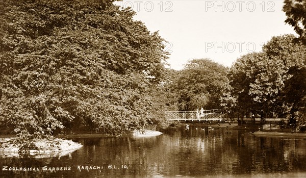 Lake in the Zoological Gardens of Karachi. A footbridge crosses an artificial lake, overhung with mature trees in the Zoological Gardens of Karachi. Karachi, India (Pakistan), circa 1910. Karachi, Sindh, Pakistan, Southern Asia, Asia.