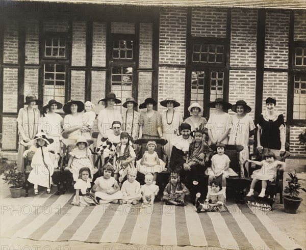 Christmas party in Burma (Myanmar). The wives and children of Public Works Department employees, pose for a group photograph at a Christmas party. The women stand at the back wearing wide-brimmed hats and dresses, whilst their children sit in the front wearing fancy dress. Two costumed men, probably entertainers, sit amongst the group. Mandalay, Burma (Myanmar), December 1924. Mandalay, Mandalay, Burma (Myanmar), South East Asia, Asia.