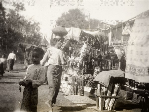 Fish festival in Shwezayan. A Burmese woman balances a ceramic pot on her head as she walks past market stalls crowded with goods at a fish festival in Shwezayan. Mandalay, Burma (Myanmar), circa February 1927. Mandalay, Mandalay, Burma (Myanmar), South East Asia, Asia.