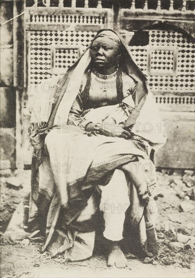 Portrait of an African woman in Egypt. Portrait of an African woman, sitting cross-legged on a chair in front of a decorative window screen. She wears a robe and head scarf, and gazes off to the left of the camera. Port Said, Egypt, circa 1920. Port Said, Port Said, Egypt, Northern Africa, Africa.