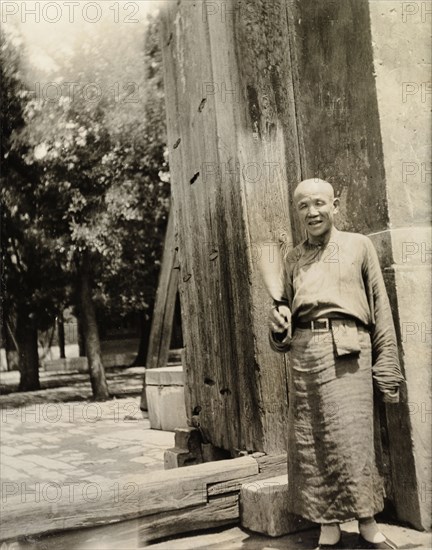 Lama priest at the doorway to a temple. A 'lama' (Buddhist monk) stands at the entrance to a temple wearing a simple robe, buckled at the waist. Peking (Beijing), China, circa 1910. Beijing, Beijing, China, People's Republic of, Eastern Asia, Asia.