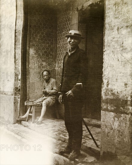 A Chinese policeman. A Chinese policeman, dressed in police uniform and a Western-style hat, smiles at the camera from a doorway. Another man sits at a workbench behind him. China, circa 1910. China, People's Republic of, Eastern Asia, Asia.