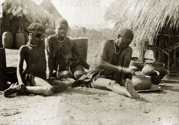 Preparing a meal. A woman and child sit on the ground outside a village hut, watching as an older woman prepares a meal in a cooking pot. Rhodesia (Zimbabwe), circa 1897. Zimbabwe, Southern Africa, Africa.