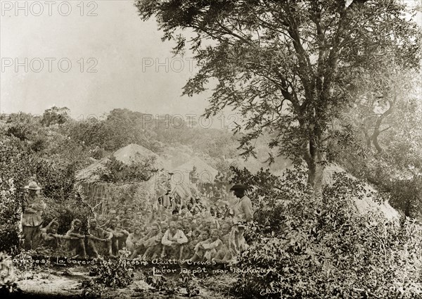 Acutt and Crewes labourers. A number of hired labourers sit on the ground in a scrubland clearing at Acutt and Crewe's labour depot near Bulawayo. They are barefoot and bareheaded, but most wear clothes of European origin. Matabeleland, Rhodesia (Matabeleland North, Zimbabwe), circa 1897., Matabeleland North, Zimbabwe, Southern Africa, Africa.