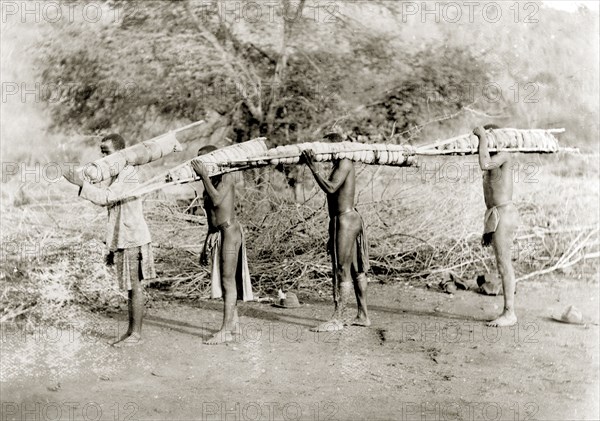 African porters, Rhodesia. Four African porters walk in line, carrying long bundles tied to poles over their shoulders. Probably Rhodesia (Zimbabwe), circa 1897. Zimbabwe, Southern Africa, Africa.
