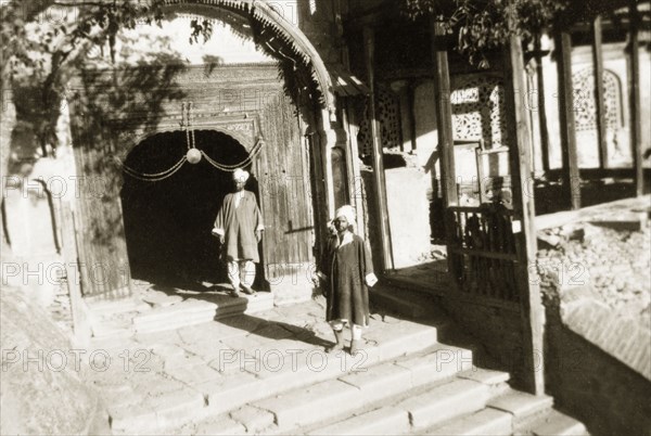 Turbaned men enter a mosque. Two Indian men stand on steps outside a doorway leading into a mosque. They are both barefoot and wear their hair up in turbans, following traditional Islamic mosque etiquette. Jammu and Kashmir State, India, circa 1927., Jammu and Kashmir, India, Southern Asia, Asia.