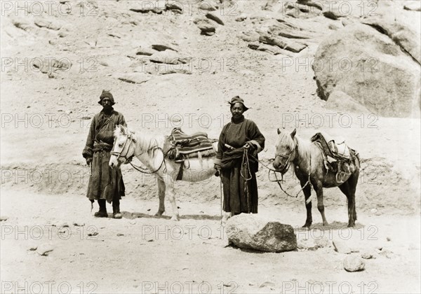 Indian men travelling with saddled ponies. Two men travel on foot across rocky terrain, accompanied by two saddled ponies, laden with blankets. Jammu and Kashmir State, India, circa 1925., Jammu and Kashmir, India, Southern Asia, Asia.