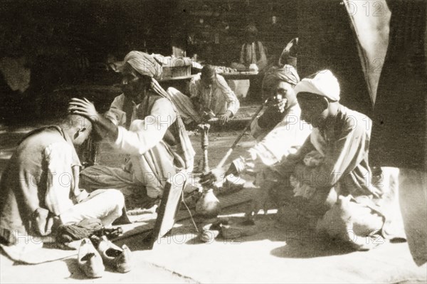 Barber's stall on a street in Peshawar. A man sits cross-legged as he has his head shaved at a barber's stall on a street in Peshawar. Two turbaned men sit nearby, one of whom smokes a 'hookah' (water pipe). Peshawar, North West Frontier Province, India (Pakistan), circa 1927. Peshawar, North West Frontier Province, Pakistan, Southern Asia, Asia.