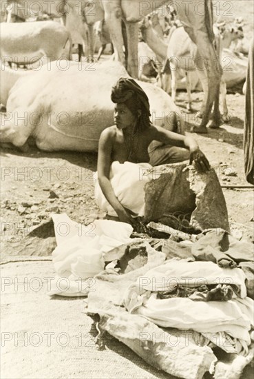 Eritrean woman setting up stall at camel market. An Eritrean woman unloads her goods, probably cloth or animal hide, onto a mat on the ground at an outdoor camel market. Eritrea, 1943. Eritrea, Eastern Africa, Africa.