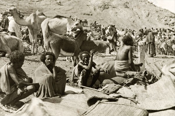 Eritrean men at a camel market. Four Eritrean men sit beside a heap of woven mats at a bustling camel market. Eritrea, 1943. Eritrea, Eastern Africa, Africa.