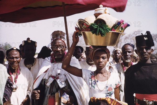 Visit of Ya Na Abdulai III. The King of Dagbon, Ya Na Abdulai III (r.1953-67), attends a celebration for the fourth anniversary of Ghanaian independence. He is preceded by a young woman (said to be a ritual water bearer) who carries ceremonial water containers in a metal tub on her head. Abdulai III wears a batakari (smock) and a hat studded with amulets for spiritual protection. Savelugu, Ghana, circa March 1961. Savelugu, North (Ghana), Ghana, Western Africa, Africa.