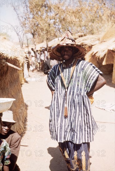 Moshi Man' at Bolgatanga market. Portrait of a man at Bolgatanga market, labelled in an original caption as 'The Moshi Man'. He wears a layered leather hat, which is typical of Bolgatanga leatherware and is commonly worn by diviners, and a smock made from blue and white striped cloth. Bolgatanga, Ghana, circa 1961. Bolgatanga, Upper East, Ghana, Western Africa, Africa.