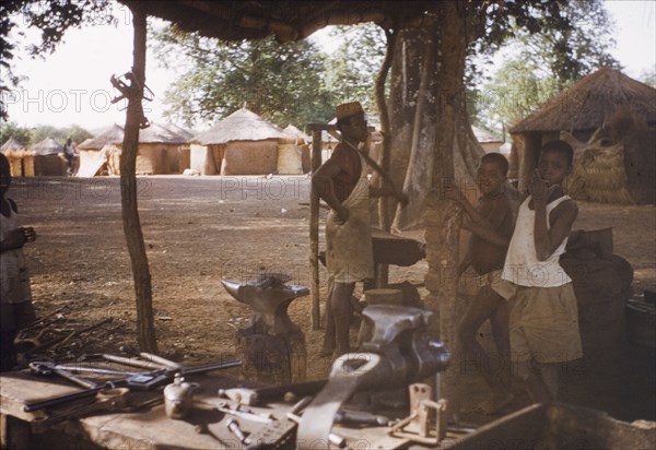 Blacksmith at Gambaga, Ghana. A blacksmith stands amongst tools in his workshop, accompanied by two young boys, possibly apprentices. Gambaga, Ghana, circa 1960. Gambaga, North (Ghana), Ghana, Western Africa, Africa.