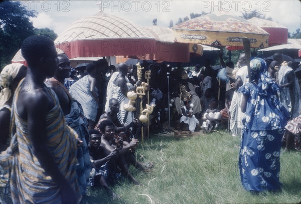 Mate Kole II at a Ngmayem festival. Mate Kole II (1910-1990), the Paramount Chief or Konor of Manya Krobo, sits beneath ceremonial umbrellas with his retinue at an annual Ngmayem harvest festival. He is attended by his 'okyeames' (linguists), each of whom carries a 'poma' (linguist's staff) as a badge of office. The child with the painted face and elaborate headdress, seated at Mate Kole II's feet, represents the Konor's 'soul'. Odumasi, Ghana, circa 1960. Odumasi, East (Ghana), Ghana, Western Africa, Africa.