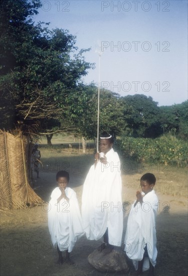 Children dressed as angels. Portrait of three children, dressed in white angel costumes for a nativity play. The two smallest children put their hands together in prayer whilst the tallest child stands on a rock, holding up a star on a stick. Ghana, circa 1960. Ghana, Western Africa, Africa.