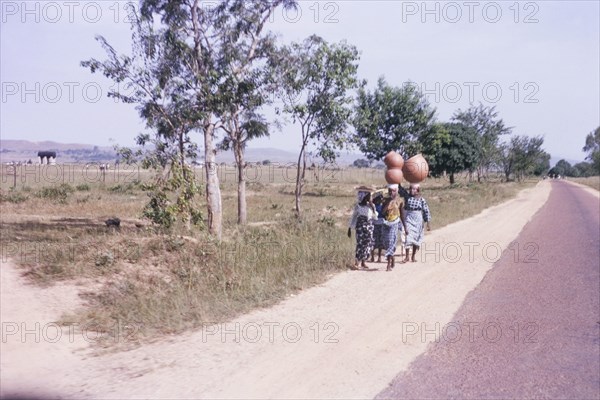 Nigerian women carrying pots. A small group of Nigerian women walk along a roadside carrying large pots on their heads. Kaduna, Nigeria, October 1963. Kaduna, Kaduna, Nigeria, Western Africa, Africa.