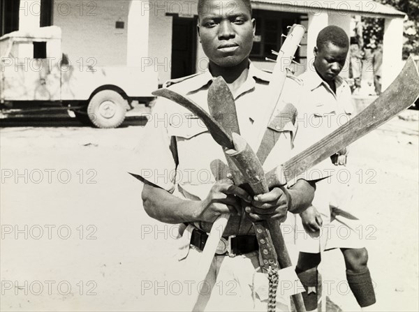 Confiscated weapons, Northern Rhodesia 1964. An officer of the Northern Rhodesia Police Force holds up a selection of weapons including pangas and machetes, which were confiscated in the violent conflict between supporters of the United National Independence Party (UNIP) and those of Alice Lenshina's Lumpa Church. Eastern Province, Northern Rhodesia (Zambia), August 1964., East (Zambia), Zambia, Southern Africa, Africa.