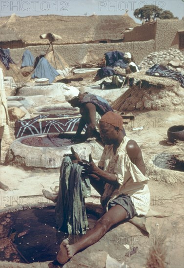 Hausa man dyeing fabric. A Hausa man sits beside a well of liquid dye, carefully dipping a section of fabric into the substance at an outdoor treatment workshop. Nigeria, circa 1975. Nigeria, Western Africa, Africa.