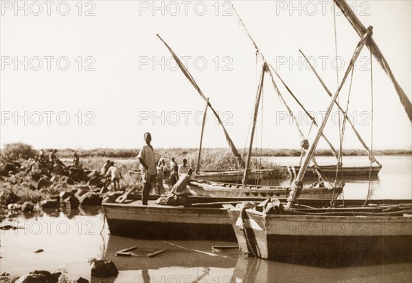 Sailboats moored in shallow water. A man stands at the stern of one of several small sailboats, which are berthed in a shallow bay at Lamu. Lamu, Kenya, 1947. Lamu, Coast, Kenya, Eastern Africa, Africa.