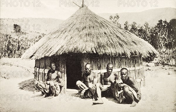 Vernacular hut in Kenya. Four men squat on the ground in front of a round hut with a thatched roof. Mombasa, British East Africa (Kenya), circa 1912. Mombasa, Coast, Kenya, Eastern Africa, Africa.