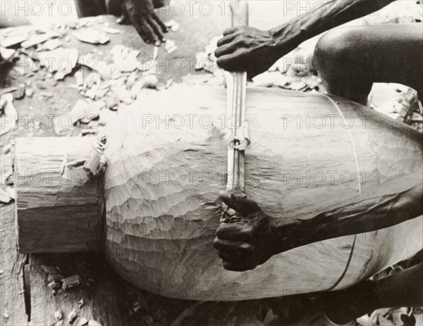 Carving the shell of an atumpan drum. A craftsman uses a long blade to carve the shell of an atumpan drum. Ghana, circa 1965. Ghana, Western Africa, Africa.