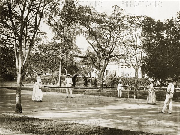 A colonial doubles match. Two British couples play doubles on a tennis court in the grounds of a large building. A turbaned Indian ball boy waits at the side of the net, ready to catch any stray shots. India, circa 1880. India, Southern Asia, Asia.