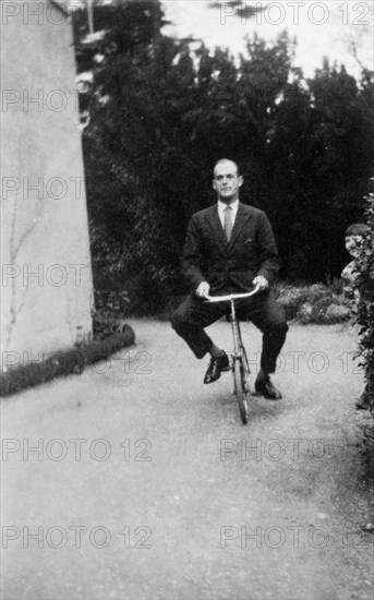 A difference of size. Courtenay Parker Bramble bends his knees awkwardly as he rides a child's bicycle down a driveway, dressed in a formal suit and tie. Gerrards Cross, Buckinghamshire, England, 16 April 1927. Gerrards Cross, Buckinghamshire, England (United Kingdom), Western Europe, Europe .