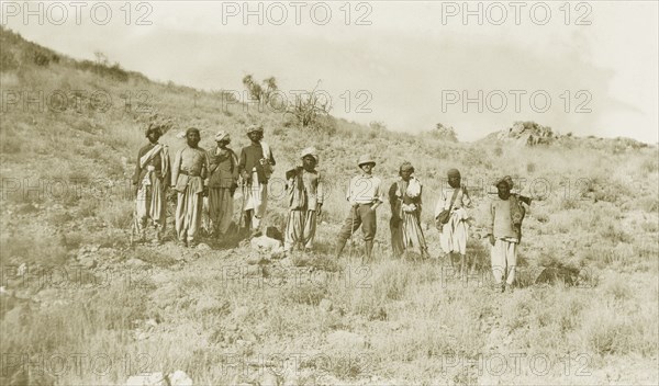 Walking sticks and a gun. Sir Henry Staveley Lawrence, Collector of Karachi, stands amongst a group of Indian men in a rocky landscape. Most of the men carry walking sticks and one of the group carries a gun slung over his shoulder. India (Pakistan), circa 1910. Pakistan, Southern Asia, Asia.